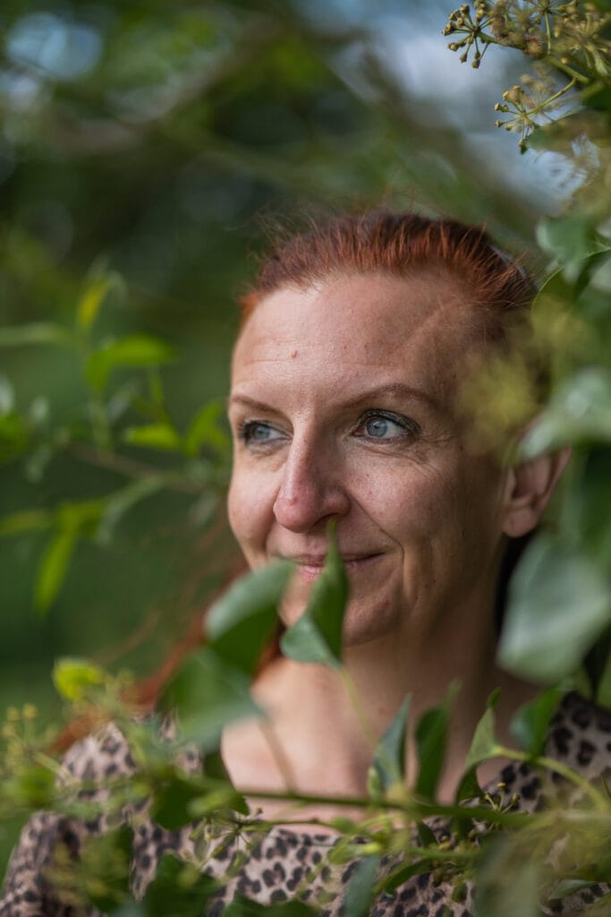 Zafir fra RestfulBlanket® ser ud gennem grønne blade, roligt smilende og omgivet af natur, symboliserer ro og bæredygtig livsstil