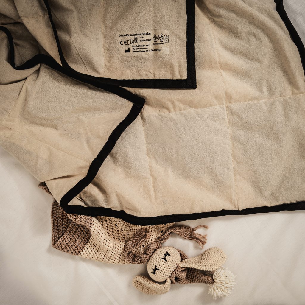 Barnets naturlige tyngdedyne fra RestfulBlanket ligger foldet på sengen sammen med et lille legetoej, som eksempel på en tryg junior tyngdedyne til børn.